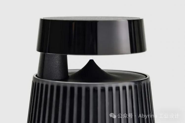 Close-up of a modern, black Bang & Olufsen wireless speaker with a cylindrical base and floating top, featuring vertical ridges on the lower section against a plain gray background.