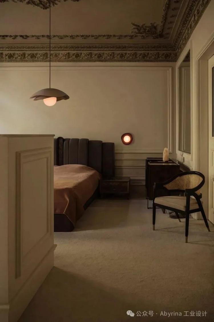 A dimly lit bedroom with a dark upholstered bed, brown bedding, a wooden desk, a chair, and ornate ceiling moldings. A pendant light and a round wall lamp provide soft lighting.