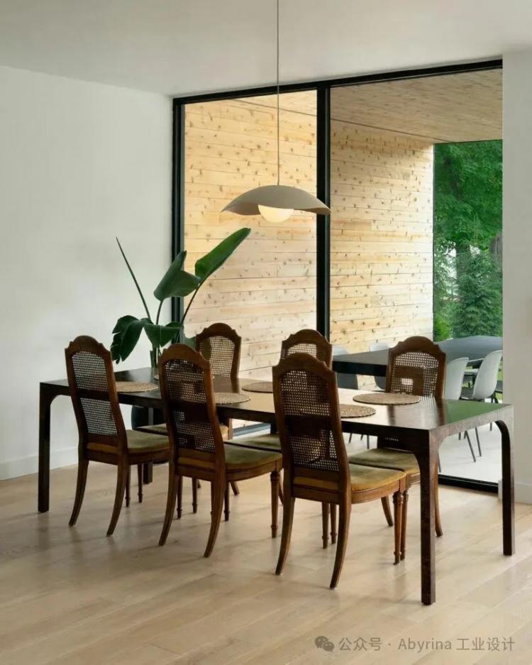 A dining room with a dark wooden table, six chairs with cane backs, a hanging pendant light, a potted plant, and large windows overlooking a wooden patio and greenery.