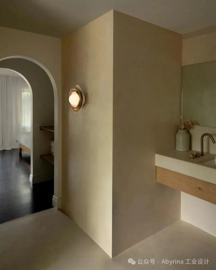 Minimalist bathroom with beige walls, a built-in sink with a wooden drawer, round wall light, large mirror, and an arched doorway leading to a bedroom with sheer curtains.