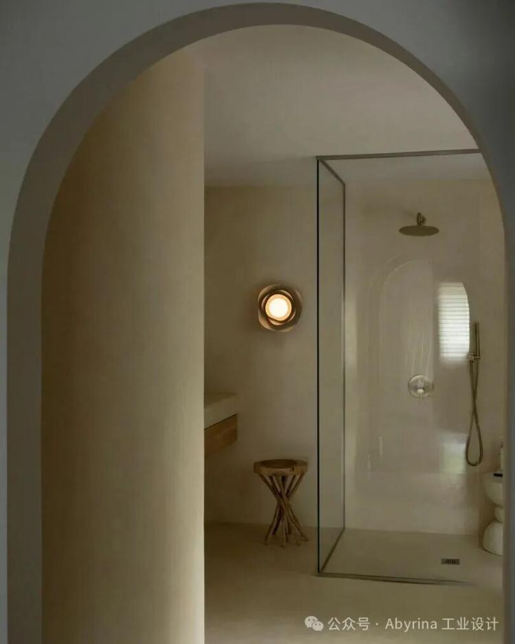 Minimalist bathroom with arched doorway, glass shower enclosure, wall-mounted showerhead, round wall light, wooden stool, and neutral-toned walls and floor.