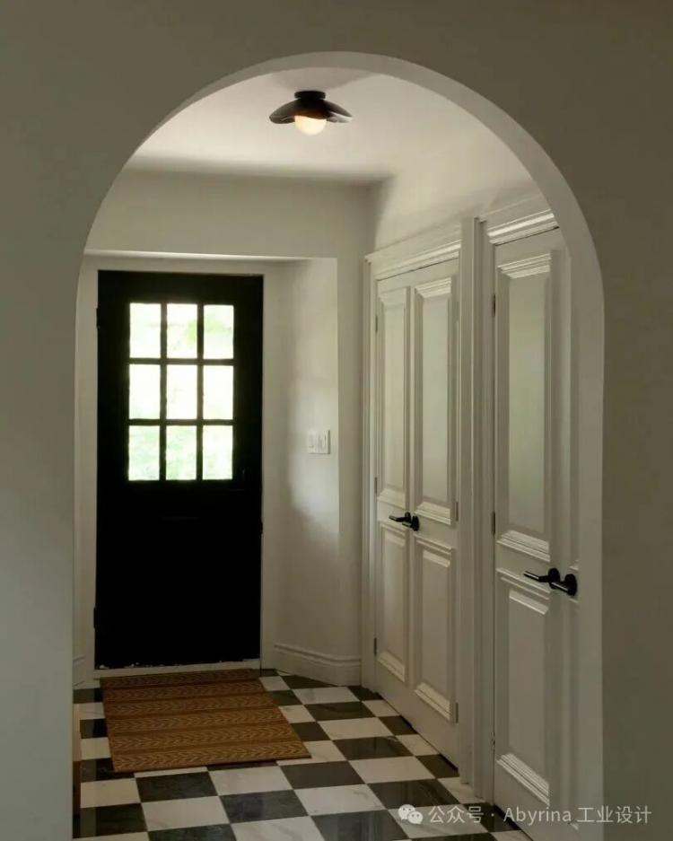 A hallway with black and white checkered floor tiles, double white doors, an arched entryway, and a black door with window panes letting in natural light.