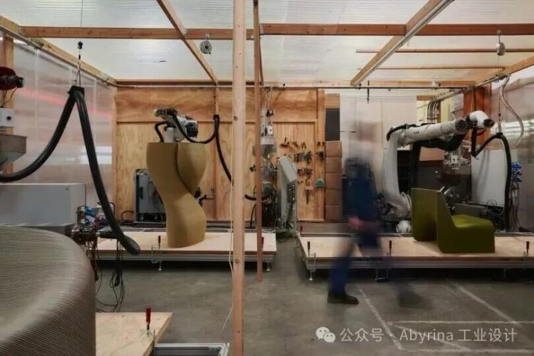 In The New Raw’s workshop, robotic arms 3D-print large sculptural objects; tools hang on the walls, and a person walks through the workspace, appearing blurred from motion.