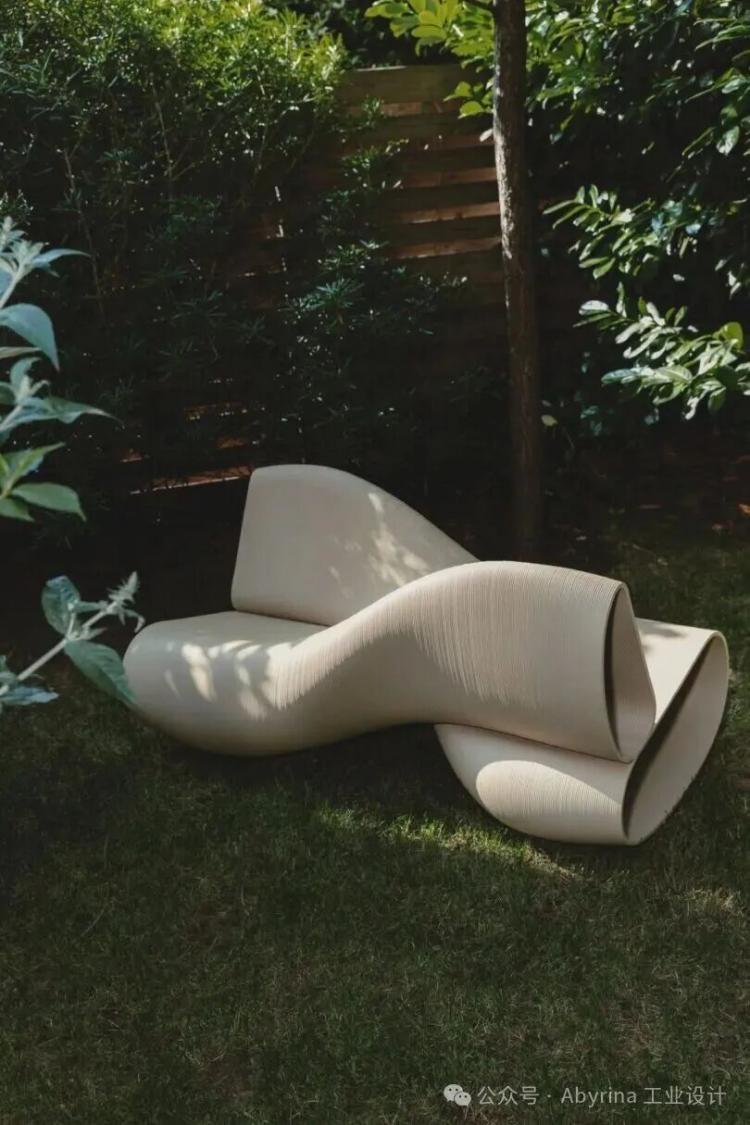 A modern, sculptural, beige lounge chair with curved lines sits on a grassy lawn surrounded by green plants and trees