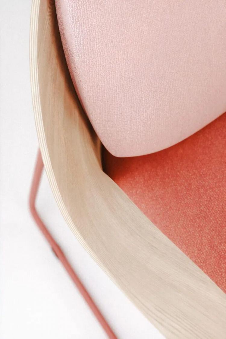 Close-up of a modern chair with a light wood frame, pink fabric backrest, and red seat cushion, featuring red metal legs on a white background.