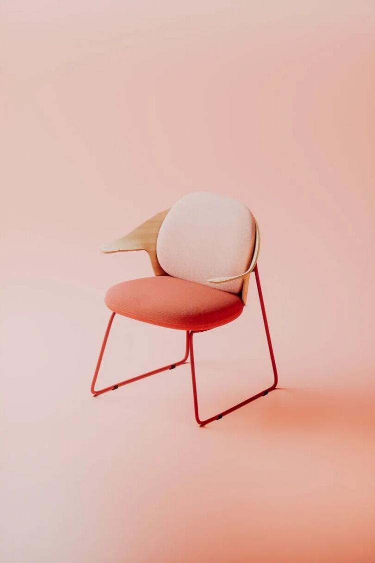 A modern chair with a light pink backrest, coral seat, wooden armrest, and red metal legs, set against a matching pale pink background.
