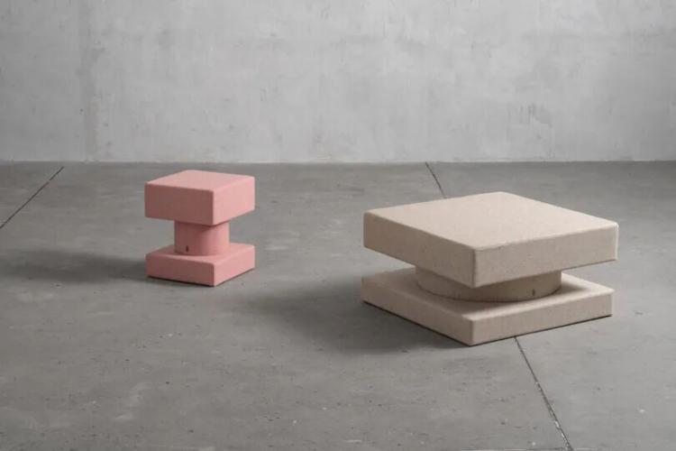 Two geometric sculptural stools or tables, one pink and one beige, placed on a gray concrete floor against a textured gray wall.