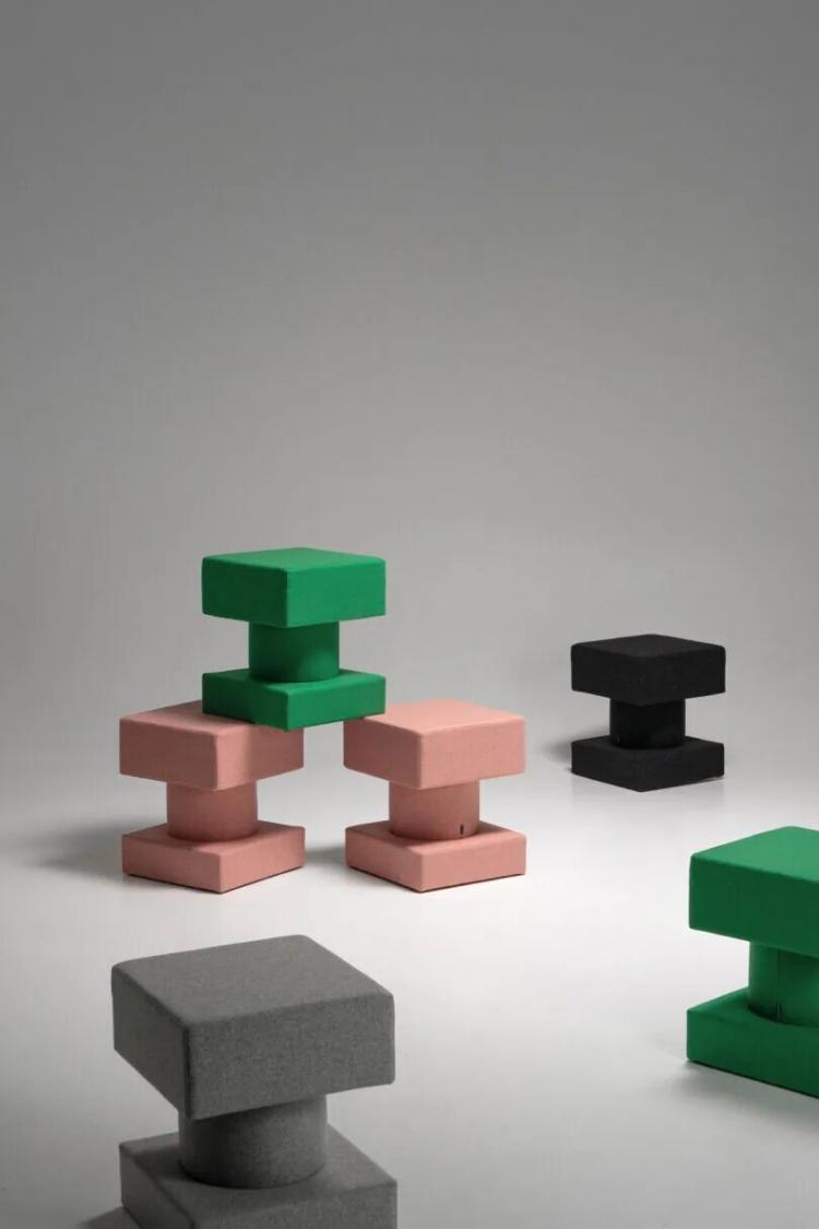 Geometric stools in green, pink, black, and gray are arranged on a plain, light-colored surface against a neutral background.