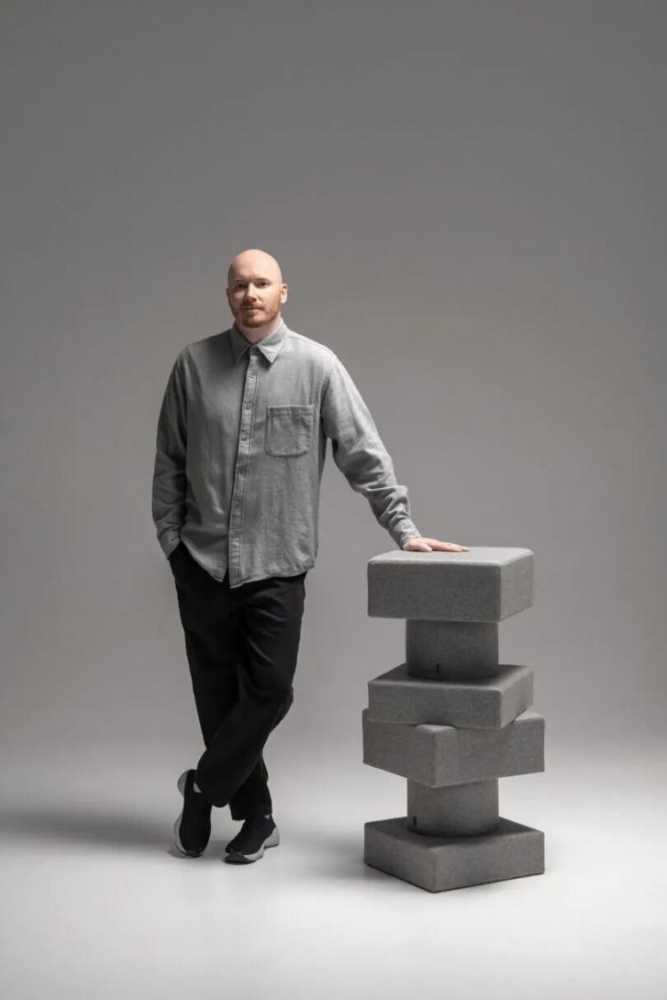 A man in a grey button-up shirt and black pants stands next to a stacked geometric sculpture made of grey blocks in a minimalist studio setting.