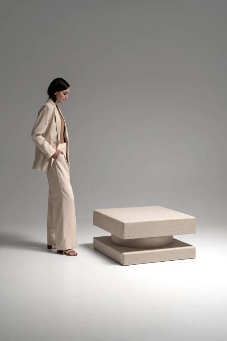A woman in a beige suit stands in a minimalist studio, looking at a modern, square, layered beige table on a white floor with a neutral background.