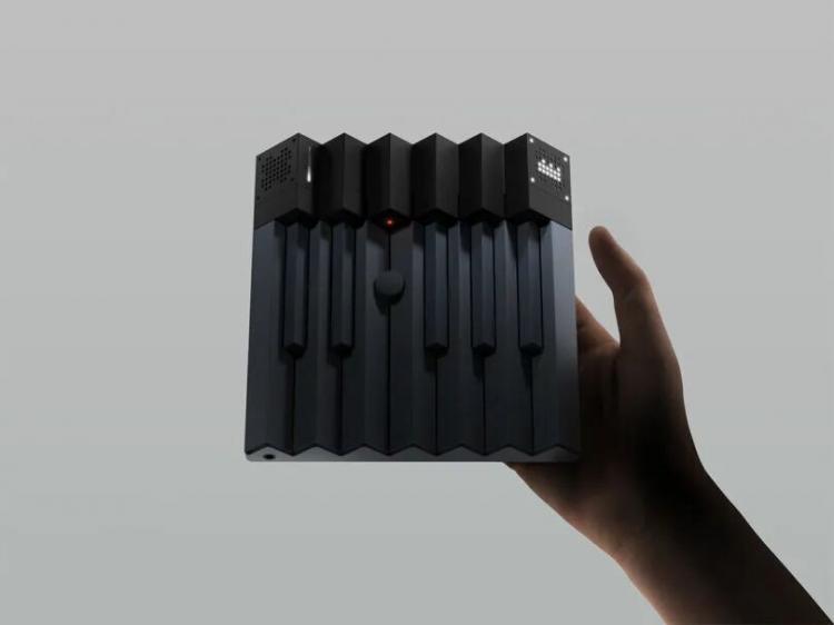 A hand holds a compact, black electronic device with piano-like keys and small LED screens on the top edge against a plain gray background.
