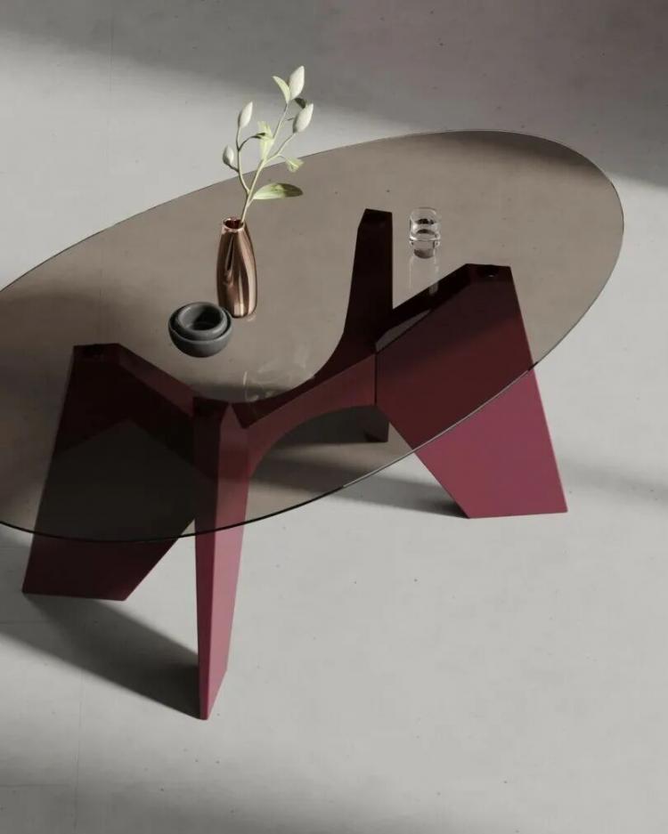 An oval glass table with a geometric maroon base, a metallic vase with a leafy branch, and a small glass placed on top, set on a light-colored floor