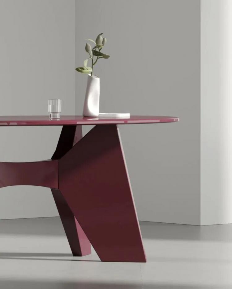 A modern maroon table with geometric legs, holding a white vase with green leaves and a clear glass of water, set in a minimalistic room with gray walls and floor