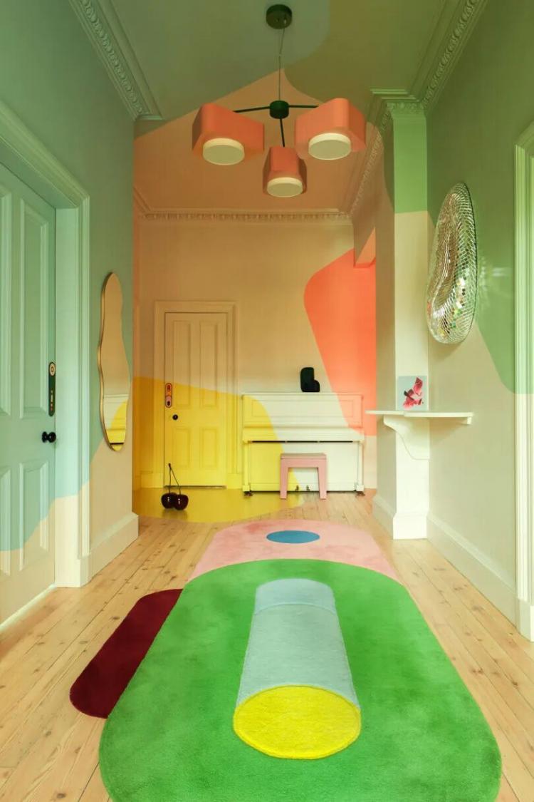 A hallway with pastel walls, geometric-patterned rug, modern light fixture, mint doors, yellow accents, and a round decorative wall mirror.