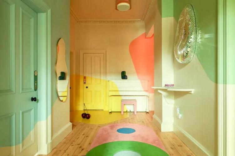 A hallway with pastel-colored walls and doors, a yellow piano, a decorative mirror, a pastel pink and green rug, and wooden floorboards.