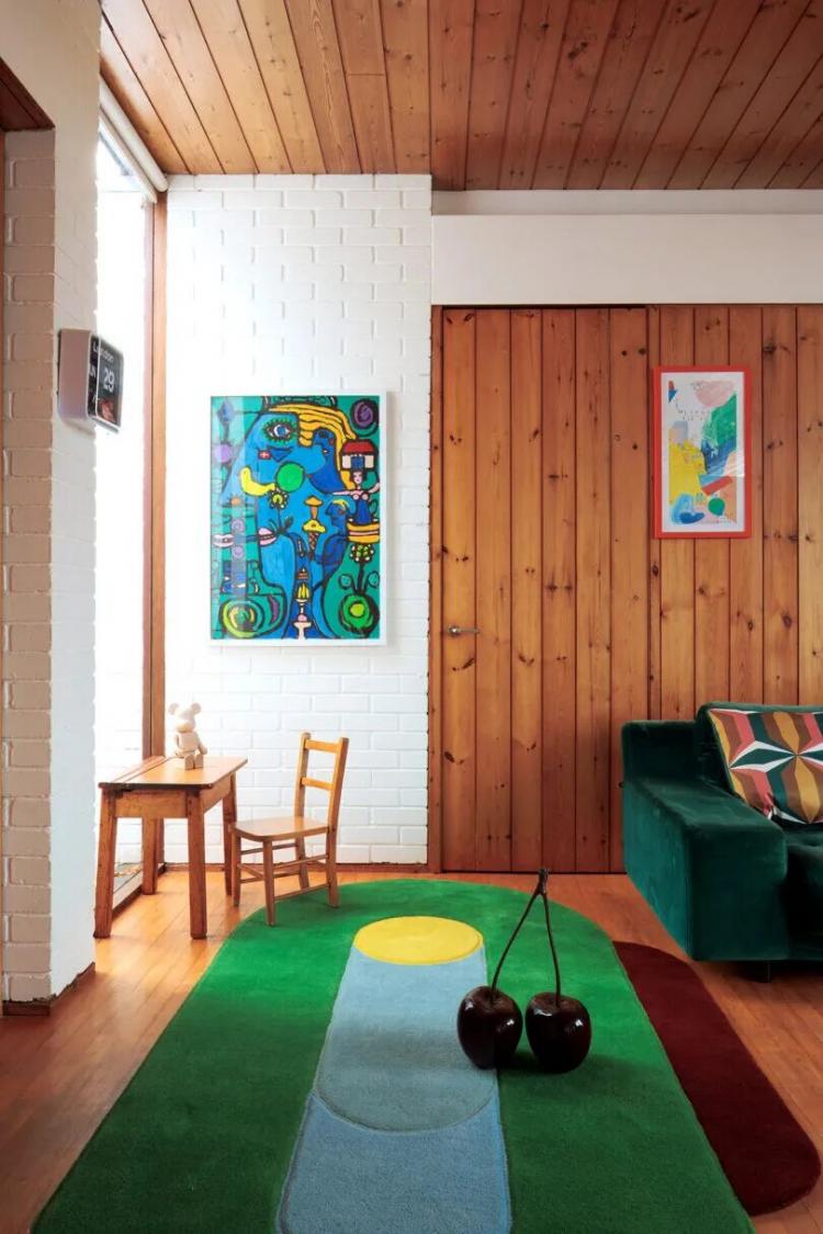 A living room with wood panel walls, a green sofa, colorful rug, small wooden table with chair, white brick wall, two artworks, and vases on the floor.