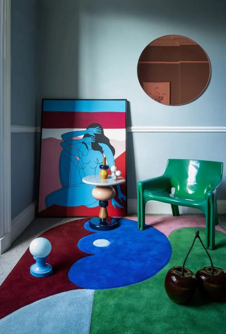A modern room with a blue and red abstract rug, green chair, round mirror, colorful art print, small table with decorative items, and two large cherry sculptures.