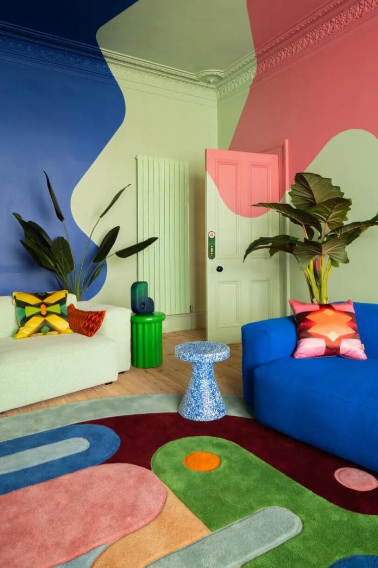 A colorful living room with abstract painted walls, bold geometric rug, blue couch, light sofa, plants, and printed pillows.
