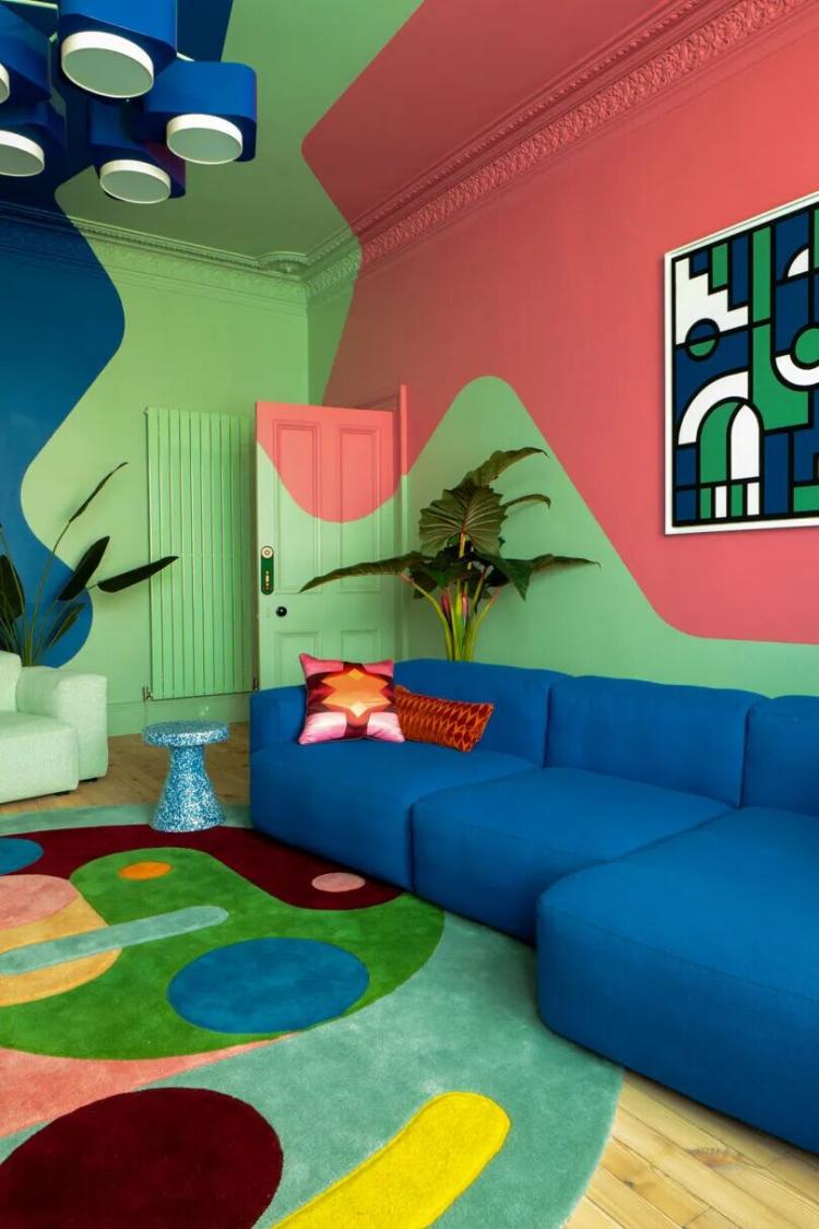 Colorful living room with bold geometric wall patterns, blue sectional sofa, graphic artwork, plants, and multicolored decor accents.