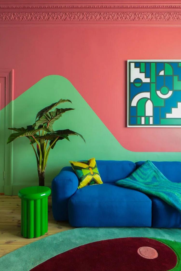 A living room with a blue couch, green side table, plant, geometric pillows, a green and pink wall, and abstract art hanging above the sofa.