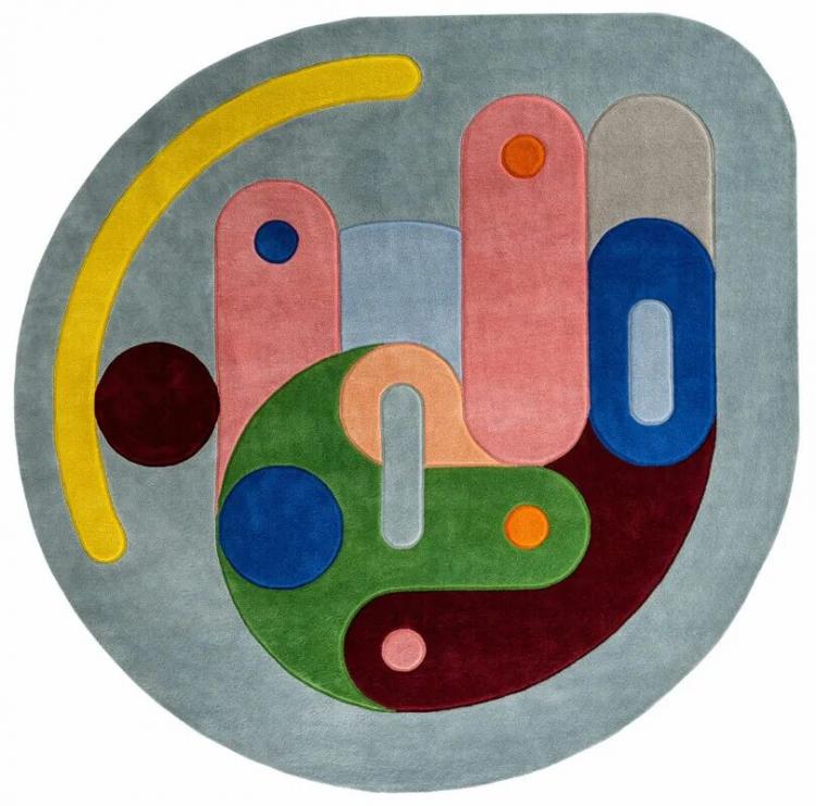 Abstract geometric rug designed by Sam Buckley, featuring overlapping shapes and vibrant colors—yellow, red, green, blue, pink, and gray—set against a light gray background.