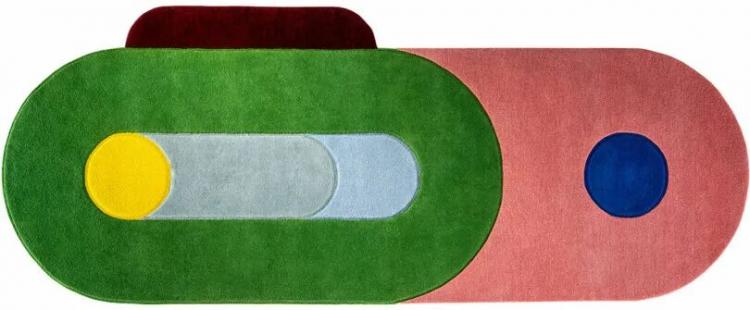 A colorful geometric rug by Sam Buckley features overlapping shapes in green, pink, red, yellow, blue, and gray tones, arranged in an eye-catching abstract pattern.
