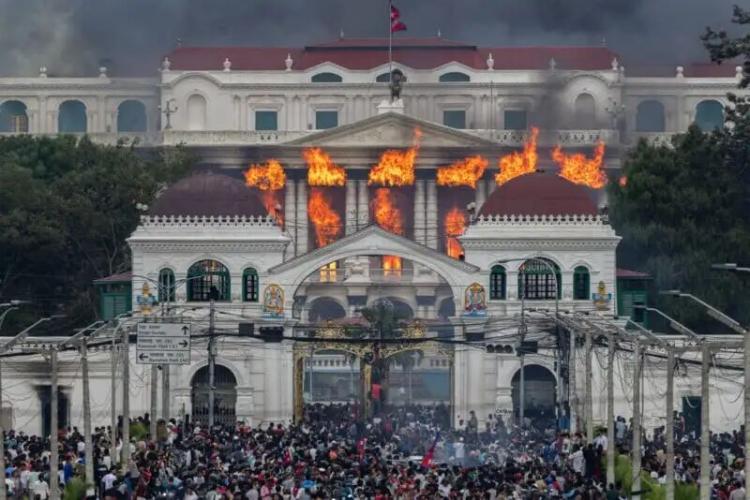 A large crowd gathers in front of a grand white building with domes as flames and smoke pour from its upper windows, indicating a major fire.