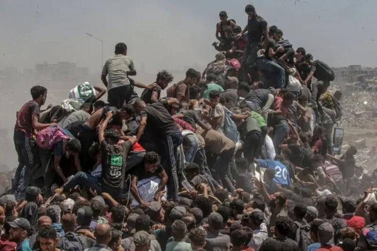 A large crowd of people climbs over each other in a chaotic, desperate scene outdoors, with dust in the air and rubble in the background, suggesting a situation of crisis or scarcity.