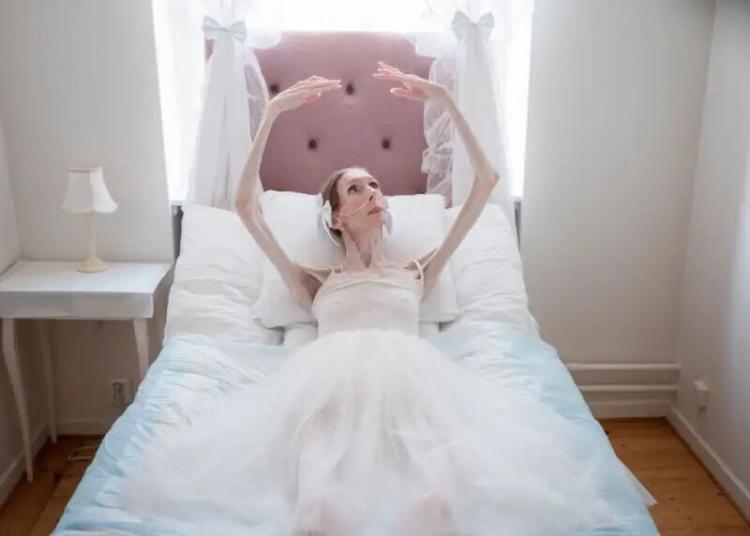 A thin woman in a white dress lies on a bed with white bedding, arms raised gracefully above her head. Sunlight streams through two windows behind her, and a small lamp sits on a bedside table.