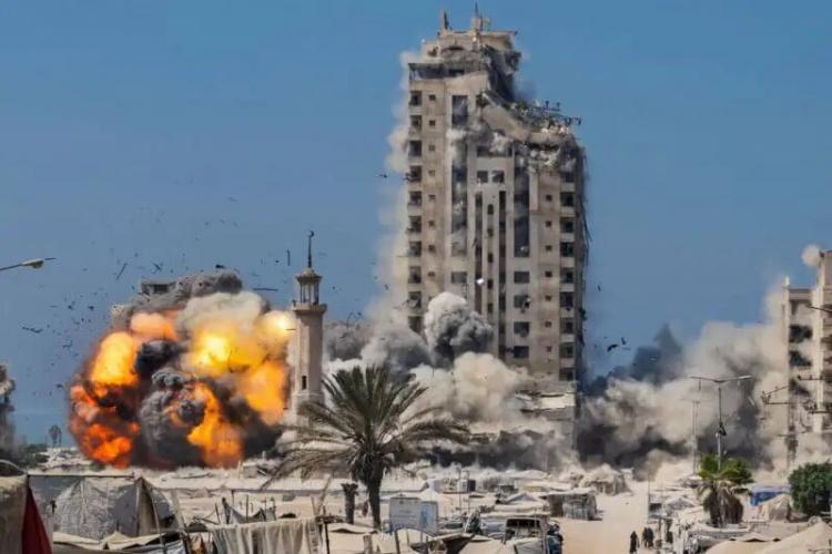 A large explosion engulfs the base of a high-rise building, sending flames, smoke, and debris into the air. The area below is filled with tents, palm trees, and power lines under a clear blue sky.